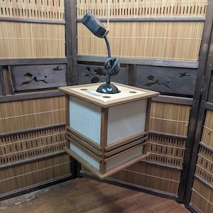 Peut inclure: Une lanterne carrée en bois avec un intérieur blanc et un bras de lampe en métal sombre et incurvé. La lanterne est suspendue à une corde et possède un cadre en bois brun clair. L'arrière-plan présente du bambou et des panneaux de bois.