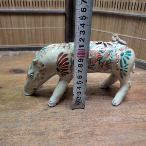 May include: A ceramic pig figurine, cream-colored with floral and leaf designs in red and teal. The figurine is approximately 4 inches tall, with a small figure on its back. The pig is on all fours, with its head lowered.