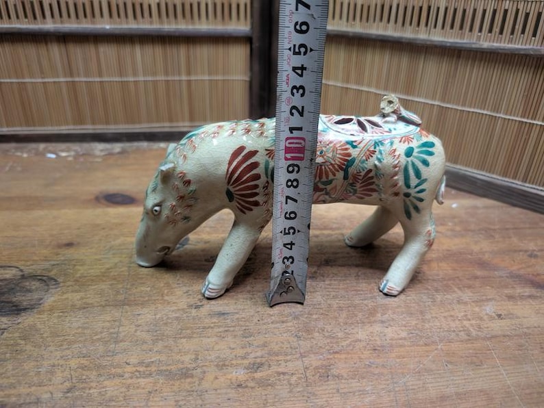 May include: A ceramic pig figurine, cream-colored with floral and leaf designs in red and teal. The figurine is approximately 4 inches tall, with a small figure on its back. The pig is on all fours, with its head lowered.