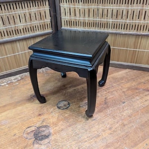 May include: A small, square, black wooden table with curved legs. The table has a flat top and a decorative edge. The table is made of wood and has a glossy finish. The table is a decorative piece and is suitable for displaying small items.