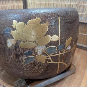 May include: A dark brown, wooden bowl with a floral design. The flower is gold with blue and white accents. The bowl has a textured surface and is set on a metal base. The bowl is likely used for decorative purposes.