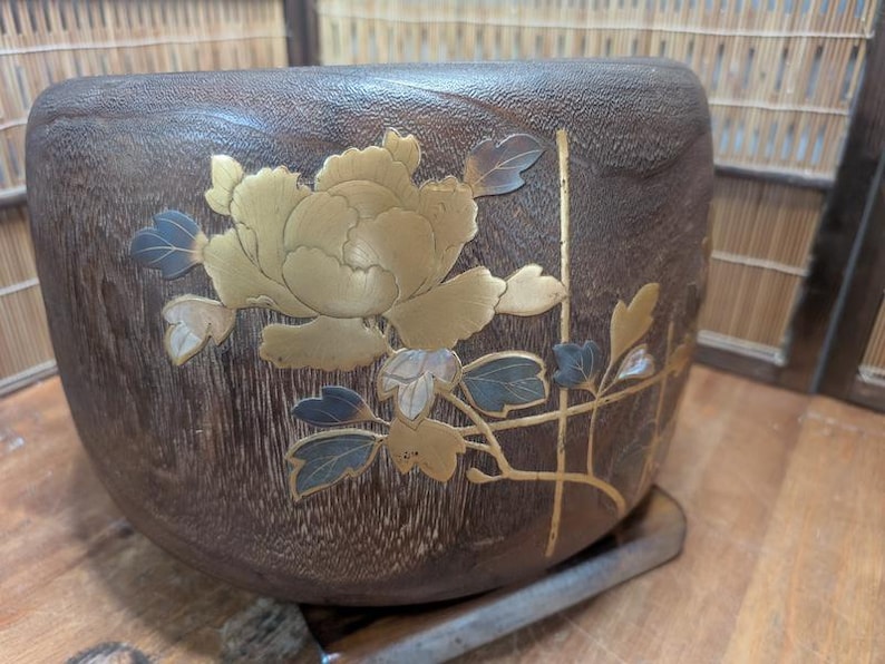 May include: A dark brown, wooden bowl with a floral design. The flower is gold with blue and white accents. The bowl has a textured surface and is set on a metal base. The bowl is likely used for decorative purposes.