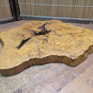 May include: A natural wood slab with a unique, irregular shape. The slab has a light brown color with darker grain patterns and a large crack running through the center. The edges are rounded and the surface is smooth.