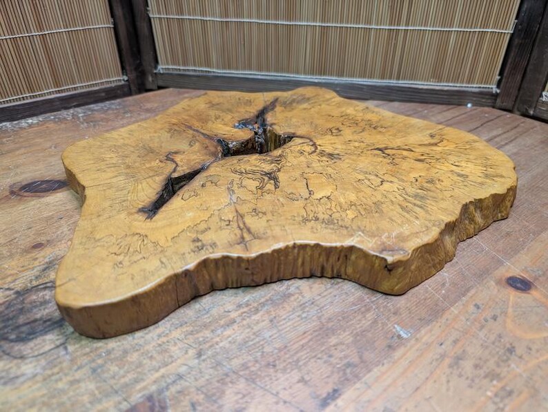 May include: A natural wood slab with a unique, irregular shape. The slab has a light brown color with darker grain patterns and a large crack running through the center. The edges are rounded and the surface is smooth.