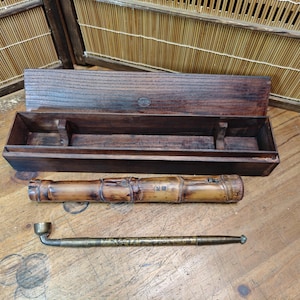 May include: An antique smoking pipe set, featuring a bamboo pipe, a metal pipe, and a wooden box. The bamboo pipe has a natural, light brown color with etched details. The metal pipe is a brass color. The wooden box is dark brown.