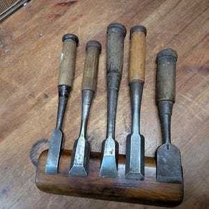 Puede incluir: Cinco cinceles de madera antiguos con mangos de madera y hojas de metal, expuestos en un soporte de madera. Los cinceles var&iacute;an en tama&ntilde;o y forma, mostrando signos de desgaste. La madera tiene un tono c&aacute;lido y natural.