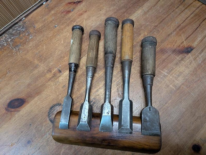 Puede incluir: Cinco cinceles de madera antiguos con mangos de madera y hojas de metal, expuestos en un soporte de madera. Los cinceles var&iacute;an en tama&ntilde;o y forma, mostrando signos de desgaste. La madera tiene un tono c&aacute;lido y natural.