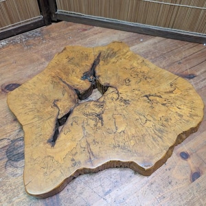 May include: A unique, irregularly shaped wooden slab with a natural edge. The slab is a warm, golden brown color with darker grain patterns and a large, irregular hole in the center. The wood appears to be unfinished and has a rustic, organic aesthetic.