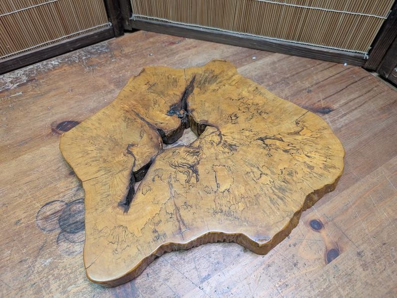 May include: A unique, irregularly shaped wooden slab with a natural edge. The slab is a warm, golden brown color with darker grain patterns and a large, irregular hole in the center. The wood appears to be unfinished and has a rustic, organic aesthetic.