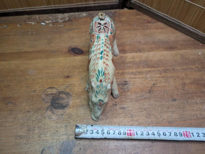 May include: A cream-colored, ceramic animal figurine with intricate floral and geometric patterns in red, green, and blue. The figurine is approximately 6 inches long, and is positioned on a wooden surface.