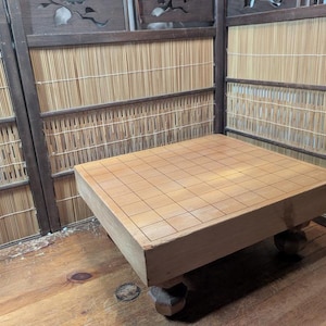 Puede incluir: Un tablero de Go de madera clara con un patrón de cuadrícula, que descansa sobre patas cortas y redondeadas. El tablero es cuadrado y parece estar hecho de madera maciza. El fondo incluye una pantalla de bambú.