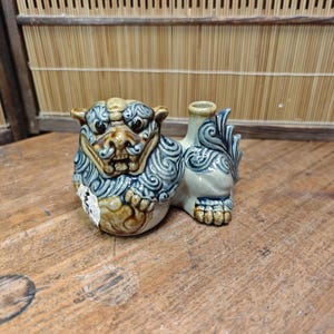 May include: A ceramic guardian lion figurine, featuring a detailed face with a fierce expression and ornate patterns in blue, brown, and cream. The figurine has a small opening on its back, suggesting it may be a small vase or incense holder.