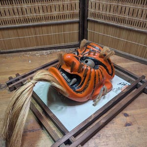 May include: An orange and black Japanese demon mask with a fierce expression, set on a wooden tray. The mask has a long, flowing mane and is displayed on a wooden surface.