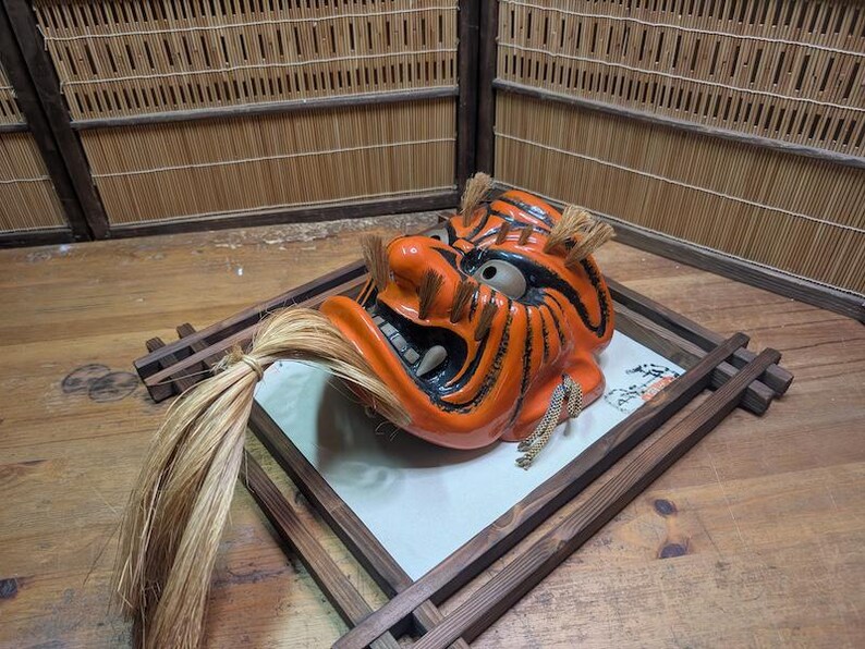 May include: An orange and black Japanese demon mask with a fierce expression, set on a wooden tray. The mask has a long, flowing mane and is displayed on a wooden surface.