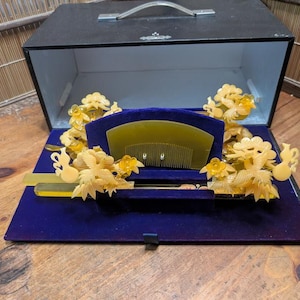 May include: A black box, open to reveal a collection of antique hair accessories. The set includes a comb and decorative ornaments in shades of yellow and gold, set against a deep blue velvet interior.