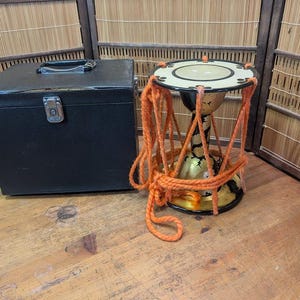 K&ouml;nnte beinhalten: Eine traditionelle japanische Trommel, oder Tsuzumi, mit einem orangefarbenen Seilgeschirr. Die Trommel hat einen schwarzen und goldenen K&ouml;rper und ein cremefarbenes Trommelfell. Links von der Trommel befindet sich ein schwarzer Koffer.