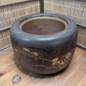 May include: A dark brown, wooden planter with a wide, rounded rim. The planter features a decorative gold floral design on its side. The interior is a lighter brown color. The planter sits on a wooden surface.