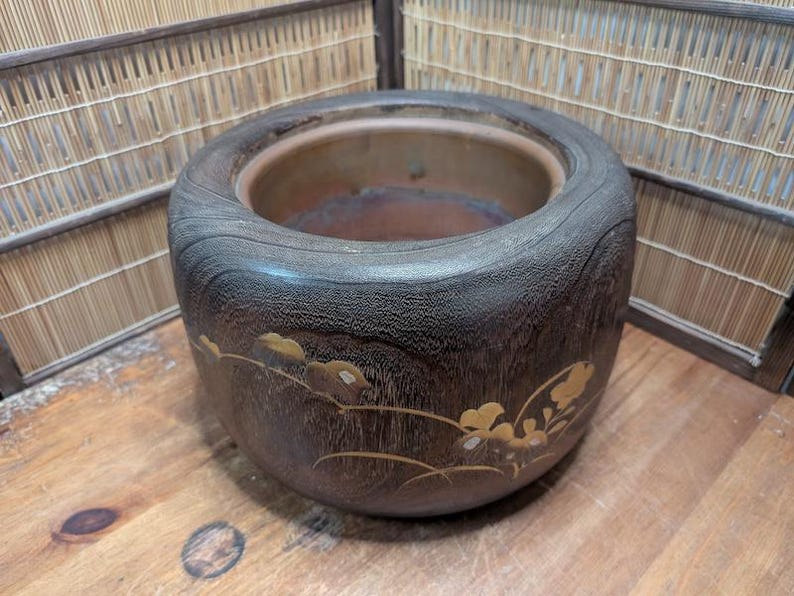 May include: A dark brown, wooden planter with a wide, rounded rim. The planter features a decorative gold floral design on its side. The interior is a lighter brown color. The planter sits on a wooden surface.
