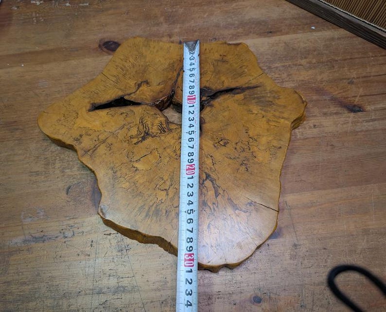 May include: A natural wood slab with a unique, irregular shape. The slab is a warm, golden brown color with darker brown and black markings. A measuring tape shows the slab is approximately 29 inches long.