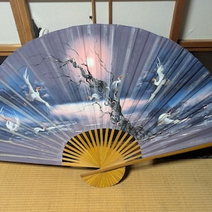 May include: Hand-painted Japanese fan depicting a sunset scene with cranes in flight. The fan has a lavender background with a tree and birds. The handle is made of natural wood.