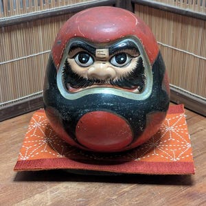 Könnte beinhalten: Dargestellt ist eine Daruma-Puppe, ein traditioneller japanischer Glücksbringer. Die Puppe hat einen roten Kopf und einen schwarzen Körper mit detailliertem Gesicht. Sie sitzt auf einem orangefarbenen und weiß gemusterten Kissen.