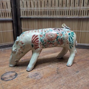 May include: A ceramic figurine of a four-legged animal, possibly a pig or hippopotamus, with a monkey on its back. The figurine is cream-colored with red and green floral patterns. The animal is on a wooden surface.