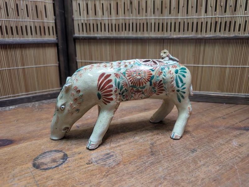 May include: A ceramic figurine of a four-legged animal, possibly a pig or hippopotamus, with a monkey on its back. The figurine is cream-colored with red and green floral patterns. The animal is on a wooden surface.