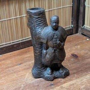 以下が含まれることがあります： 彫刻された木の幹のデザインの濃い茶色のセラミック花瓶。詳細な戦士の姿が花瓶の隣に立っています。戦士は詳細な衣装を着ています。花瓶とフィギュアは木製の表面にあります。