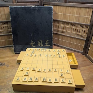 May include: A yellow and black Japanese Shogi board game set. The set includes a wooden game board with grid lines, game pieces, a black box with gold Japanese characters, and a drawer for storing pieces.