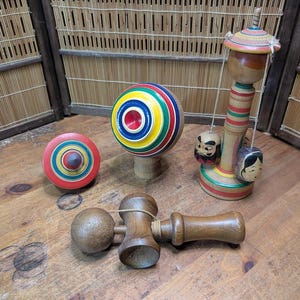 May include: A collection of traditional Japanese wooden toys, including spinning tops and a kendama. The tops feature vibrant colors like red, yellow, and blue. The kendama is a wooden toy with a ball and a handle.