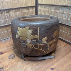 May include: A dark brown, round wooden planter with a floral design. The planter features a large, gold-colored peony with blue and gold accents. It sits on a small, square wooden base.