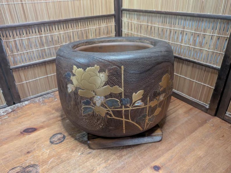 May include: A dark brown, round wooden planter with a floral design. The planter features a large, gold-colored peony with blue and gold accents. It sits on a small, square wooden base.