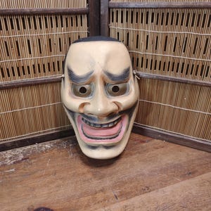 May include: A vintage Japanese Noh theater mask with a smiling expression. The mask is painted in beige, with black eyebrows and a black hairline. The mouth is open, revealing pink lips and teeth. The mask is displayed against a bamboo screen.