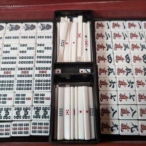 Op de afbeelding: Een complete Mahjong-set in een rode koffer. De set bevat witte tegels met zwarte en rode markeringen, dobbelstenen en stokjes. De tegels zijn in compartimenten in de koffer geplaatst.