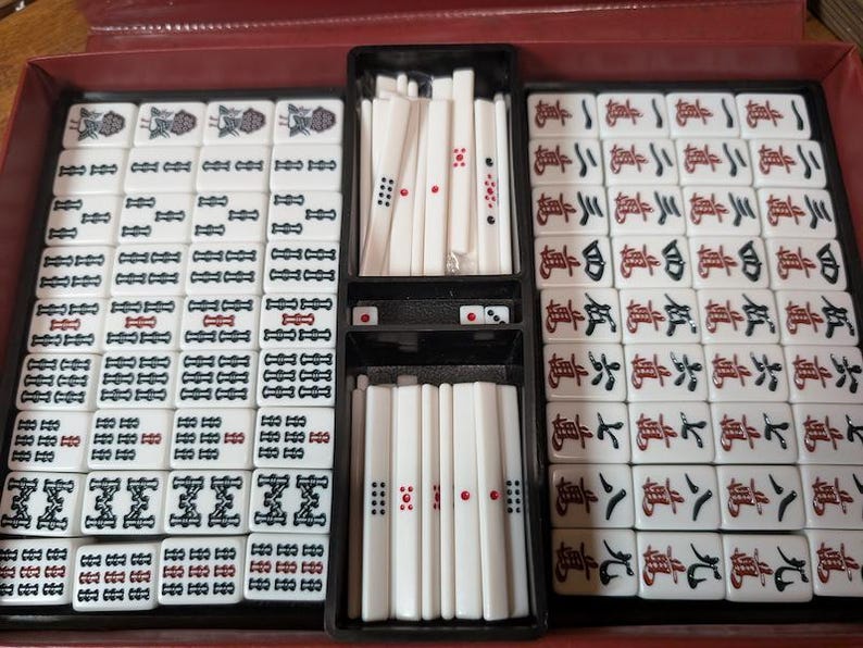 Op de afbeelding: Een complete Mahjong-set in een rode koffer. De set bevat witte tegels met zwarte en rode markeringen, dobbelstenen en stokjes. De tegels zijn in compartimenten in de koffer geplaatst.
