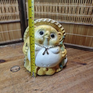 May include: A ceramic tanuki statue with a straw hat, black eyes, and a white belly. The statue is approximately 7 inches tall. It has a chain around its neck and is holding a leaf. The statue is on a wooden surface.