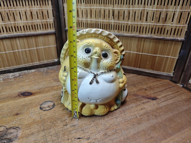 May include: A ceramic tanuki statue with a straw hat, black eyes, and a white belly. The statue is approximately 7 inches tall. It has a chain around its neck and is holding a leaf. The statue is on a wooden surface.