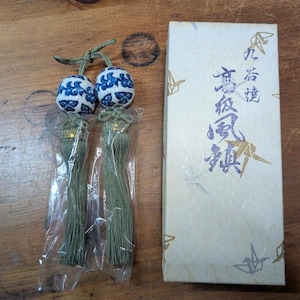 May include: Two ceramic spheres featuring a blue and white floral pattern, each attached to a long olive green tassel. The spheres are partially encased in clear plastic packaging. Beside them is a rectangular off-white box with gold crane illustrations and blue Japanese calligraphy text.