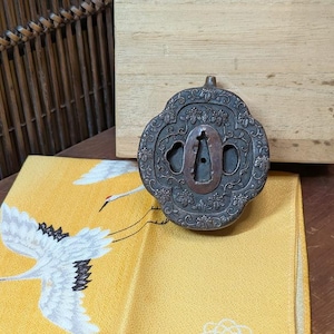 May include: A detailed, antique metal Japanese tsuba, a sword guard, with intricate floral and geometric patterns. The tsuba is dark gray with a bronze-colored center and sits on a yellow fabric with crane illustrations. A wooden box is in the background.