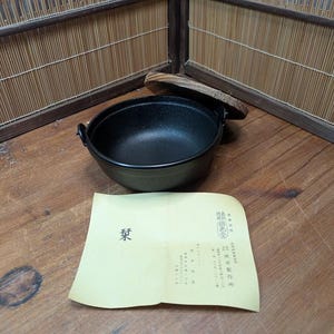Op de afbeelding: Een zwarte gietijzeren pot met twee handvatten en een houten deksel. Een geel papier met Japanse tekst ligt voor de pot. De pot staat op een houten oppervlak.
