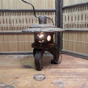 May include: A dark brown wooden lantern with a unique design. The lantern has a circular top and a base with three legs. The body of the lantern features cut-out shapes that allow light to shine through. The lantern is lit, creating a warm glow.