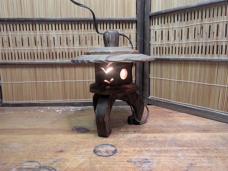 May include: A dark brown wooden lantern with a unique design. The lantern has a circular top and a base with three legs. The body of the lantern features cut-out shapes that allow light to shine through. The lantern is lit, creating a warm glow.