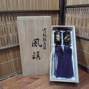 May include: A wooden box with Japanese characters and a decorative set of black and gold tassels with purple fringe. The tassels are displayed in a white box. The background is a bamboo screen.