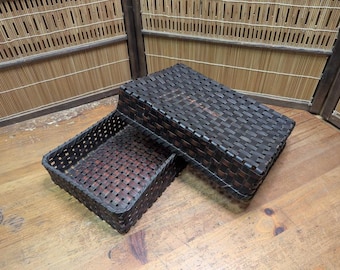 Puede incluir: Una cesta tejida de color marrón oscuro con una tapa a juego. La cesta es rectangular con un patrón tejido y está abierta. La tapa también es rectangular y encaja sobre la cesta. La cesta y la tapa están hechas de un material marrón oscuro.