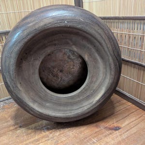 May include: A large, dark brown wooden bowl with a wide, rounded rim and a circular opening in the center. The bowl has a smooth, polished surface and appears to be handmade. The bowl is sitting on a wooden surface.