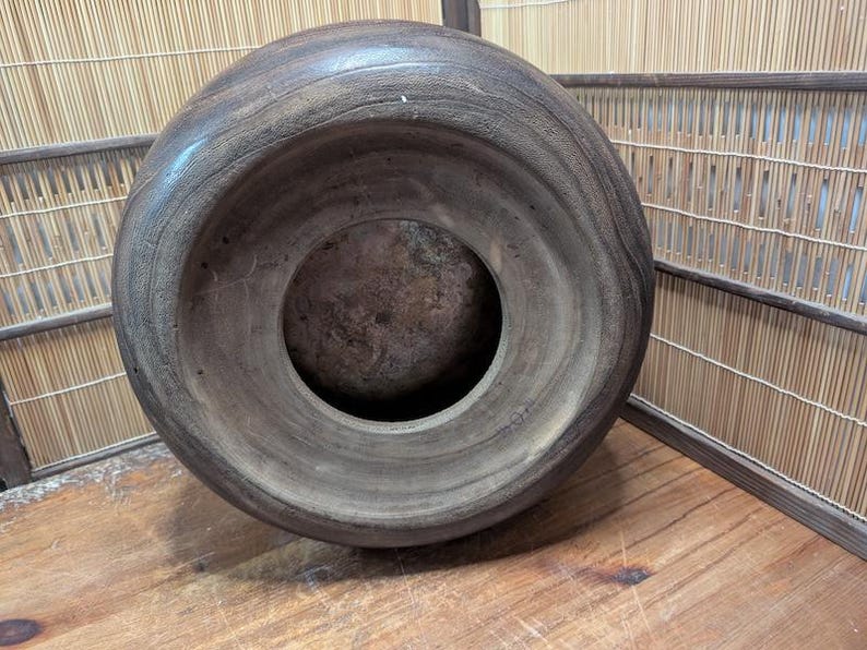 May include: A large, dark brown wooden bowl with a wide, rounded rim and a circular opening in the center. The bowl has a smooth, polished surface and appears to be handmade. The bowl is sitting on a wooden surface.