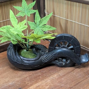 Op de afbeelding: Een zwarte, gesneden houten plantenbak in de vorm van een waterrad met groene kunstplanten. De plantenbak heeft een gedetailleerd ontwerp en staat op een houten oppervlak. De bladeren zijn heldergroen.