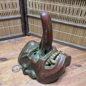 Op de afbeelding: Een vintage, handgeschilderd Japans masker in rood en groen met gouden tanden, met een lange, gebogen neus. Het masker ligt op een houten oppervlak, met een bamboe scherm op de achtergrond. Uniek decoratief item.