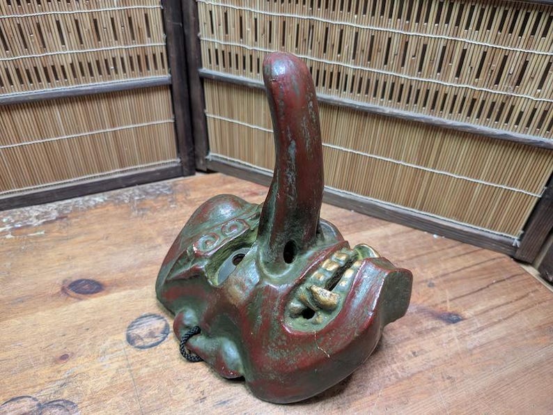 Op de afbeelding: Een vintage, handgeschilderd Japans masker in rood en groen met gouden tanden, met een lange, gebogen neus. Het masker ligt op een houten oppervlak, met een bamboe scherm op de achtergrond. Uniek decoratief item.