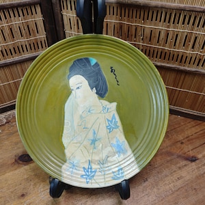 May include: A round, olive-green ceramic plate featuring a Japanese geisha design. The geisha is depicted in a kimono with blue leaf patterns. The plate is supported by a black stand and has a ridged edge.
